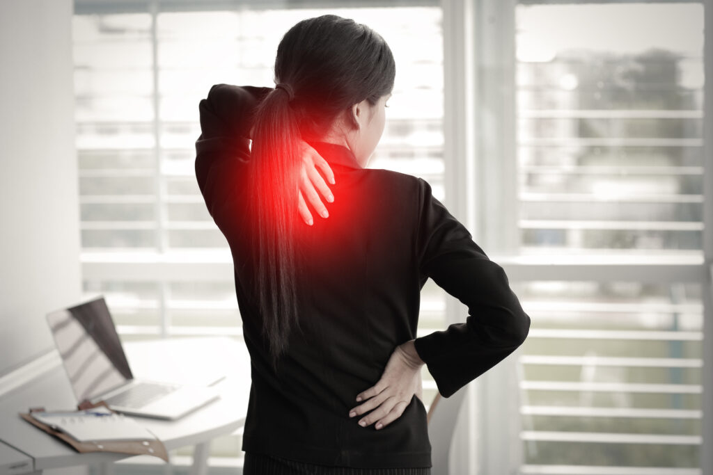 Professional woman in black blazer experiencing neck and lower back pain while standing in an office, with red highlighted pain points indicating physical strain from overwork