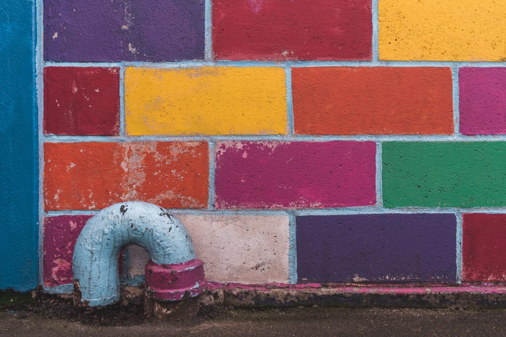 Weathered pipe elbow mounted at the base of a vibrant multicolored brick wall, showing how functional constraints exist within—and against—a backdrop of color and dimension