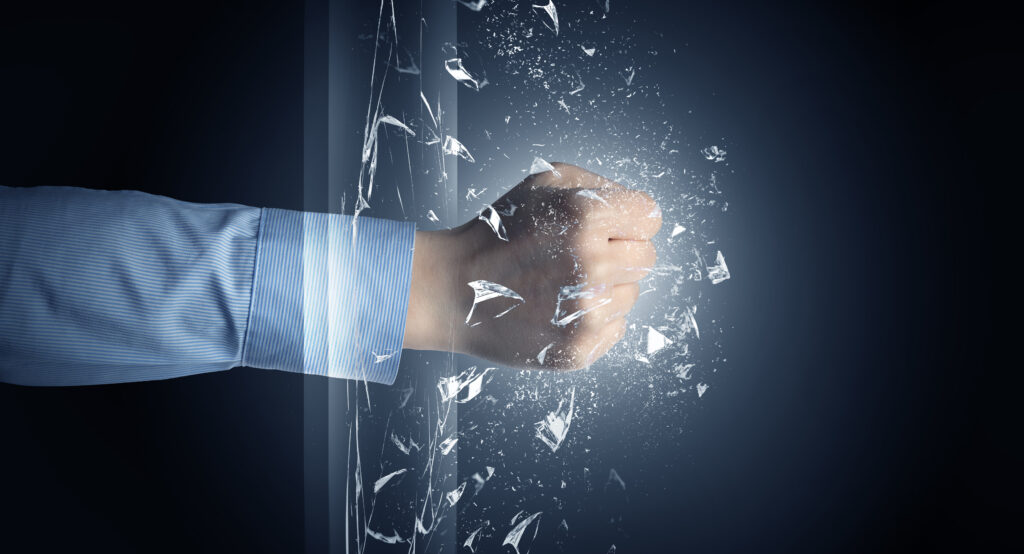 A fist in a blue dress shirt sleeve punching through a glass pane, shattering it into fragments against a dark blue background. Used as the featured image for H2P Collective blog post on breaking through barriers.