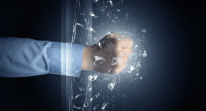 A fist in a blue dress shirt sleeve punching through a glass pane, shattering it into fragments against a dark blue background. Used as the featured image for H2P Collective blog post on breaking through barriers.