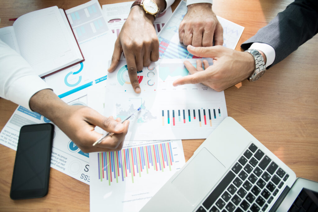 Overhead view of three people's hands pointing at and reviewing a spread of colorful charts, graphs, and data reports on a wooden conference table, with a laptop and smartphone visible alongside the printed analytics.