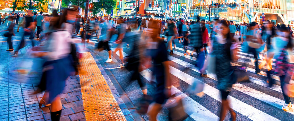 Long-exposure photograph of crowds crossing the Shibuya scramble intersection in Tokyo at night, figures motion-blurred in multiple directions against glowing neon storefronts, capturing the simultaneous movement of dozens of people navigating the same space on their own timelines.