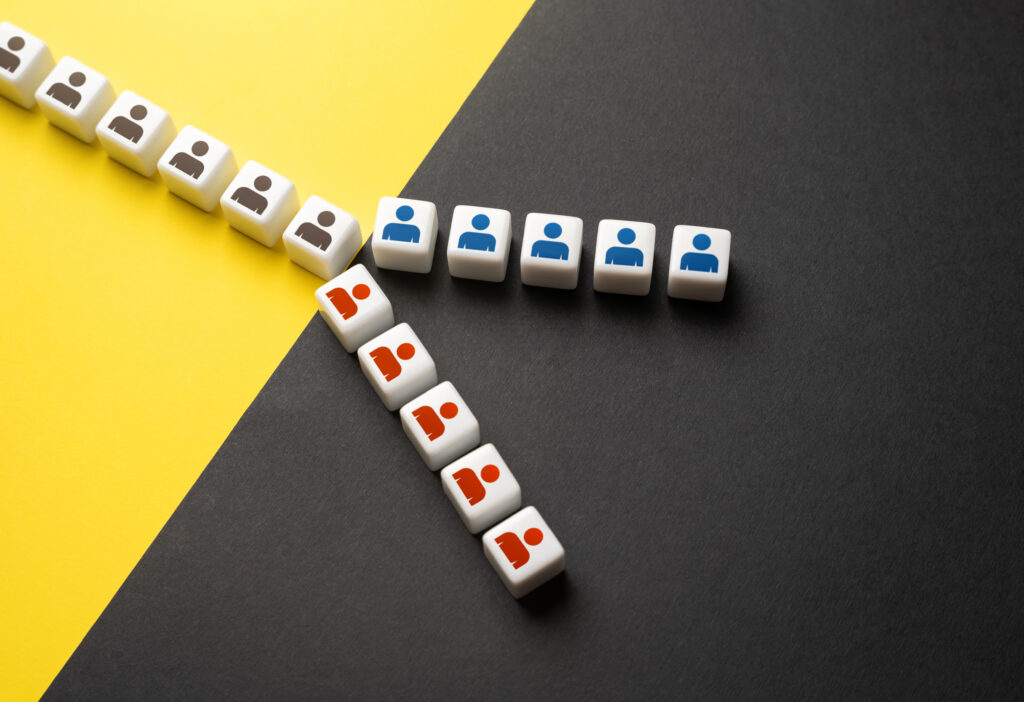White dice cubes printed with person icons—gray, blue, and red/orange—arranged in a forking pattern against a split yellow and dark gray background, representing a group divided into distinct segments moving in different directions.