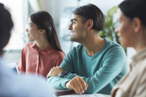 Young professional in teal sweater sits with colleagues at a meeting table, listening attentively as someone speaks off-camera, demonstrating the practice of receiving guidance without prescribing direction.