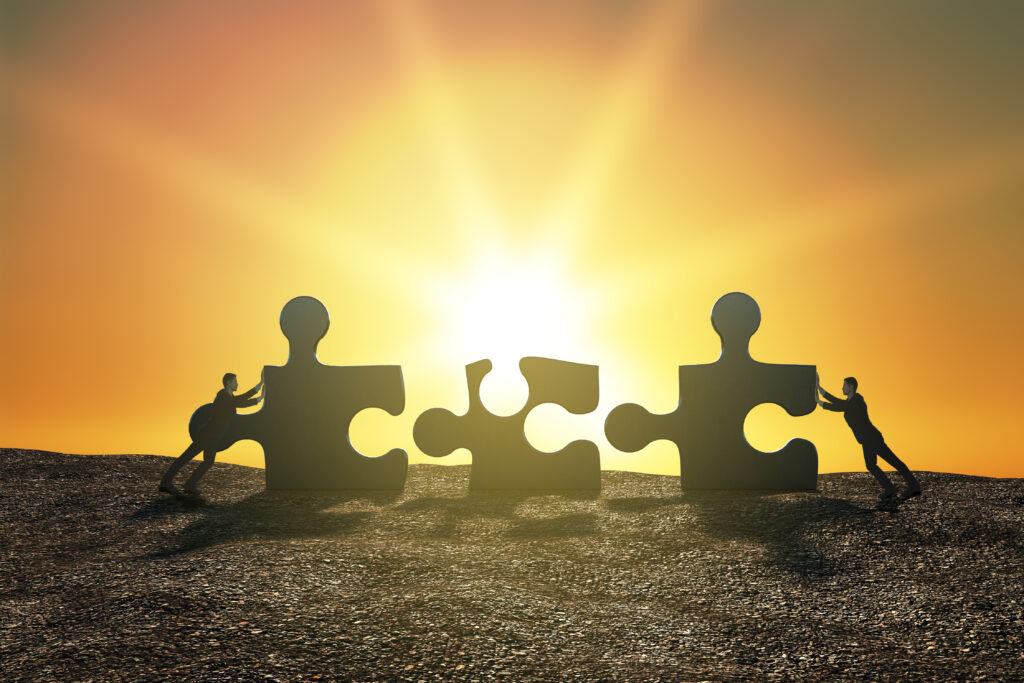 Silhouetted figures push large puzzle pieces toward one another against a sunrise sky, representing the deliberate construction of legacy through aligned effort that outlasts any single contributor.
