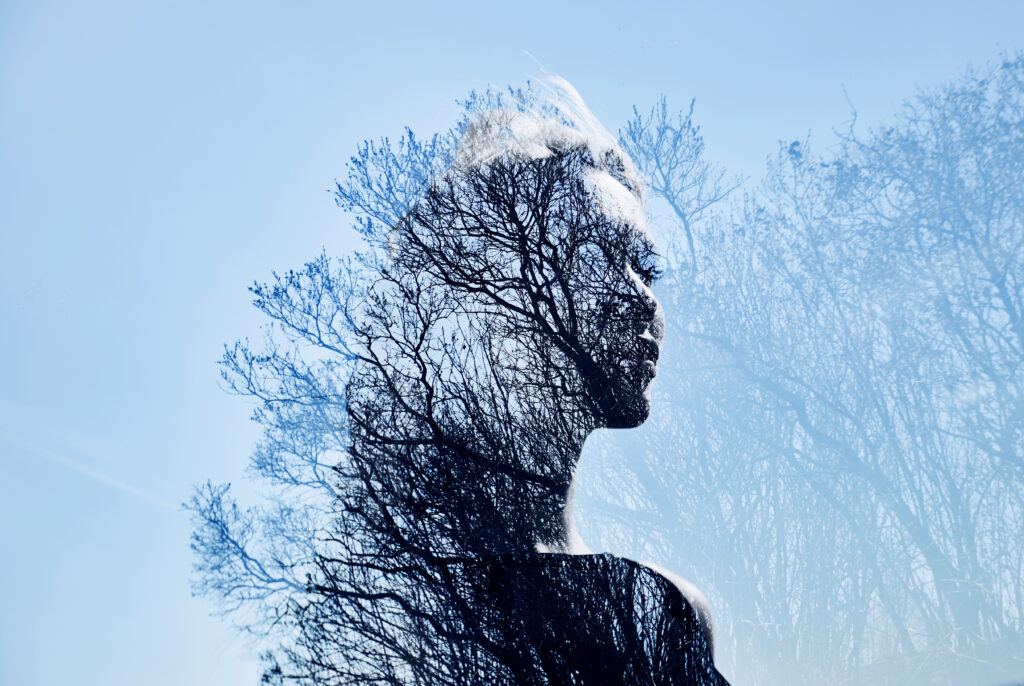 Double-exposure profile of a person merged with bare tree branches against a pale blue sky, illustrating identity as something that emerges organically through what's been weathered, not something discovered separately from the experience.