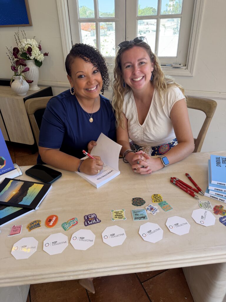 Liz St. James signs a copy of Unrestricted for a smiling attendee at a book-signing table set with copies of Synovial Space and H2P Collective materials