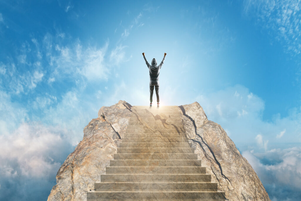 Person in a gray hoodie standing at the top of a stone staircase on a mountain peak, arms raised above the clouds.