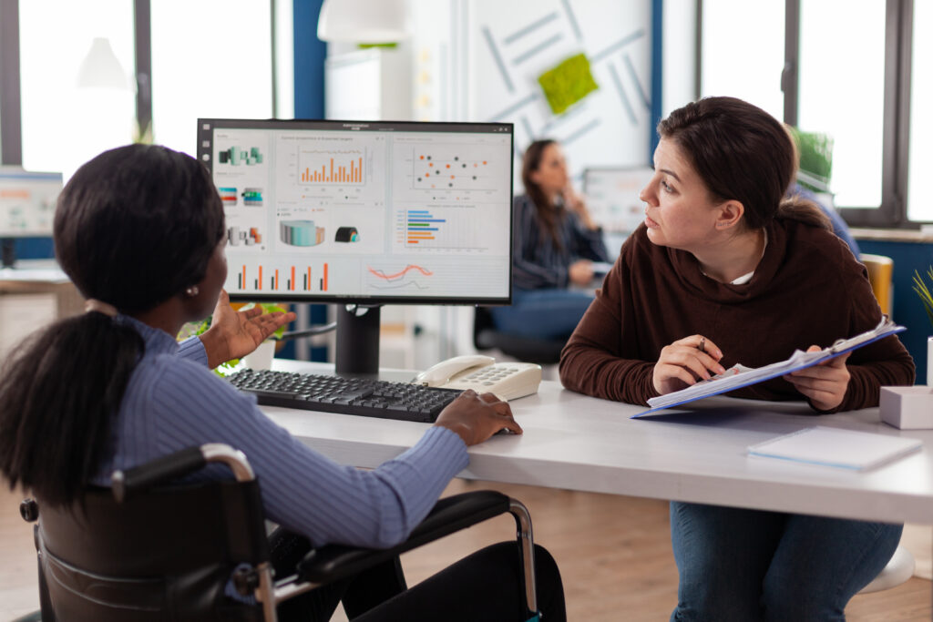 Two colleagues review analytics dashboards displayed on a monitor at an office desk, one seated in a wheelchair using the keyboard and the other seated across with notes in hand, engaged in focused review of the metrics on screen.