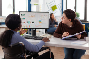 Two colleagues review analytics dashboards displayed on a monitor at an office desk, one seated in a wheelchair using the keyboard and the other seated across with notes in hand, engaged in focused review of the metrics on screen.