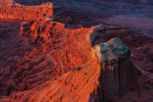 Aerial view of red sandstone canyon formations in Utah's Canyonlands region at sunset, with a narrow winding trail cutting along the ridgeline of a large mesa, warm golden light illuminating the layered rock.