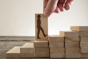 A hand lowers a wooden block carved with the silhouette of a walking man onto the next stair in a rising staircase built from stacked wooden blocks.