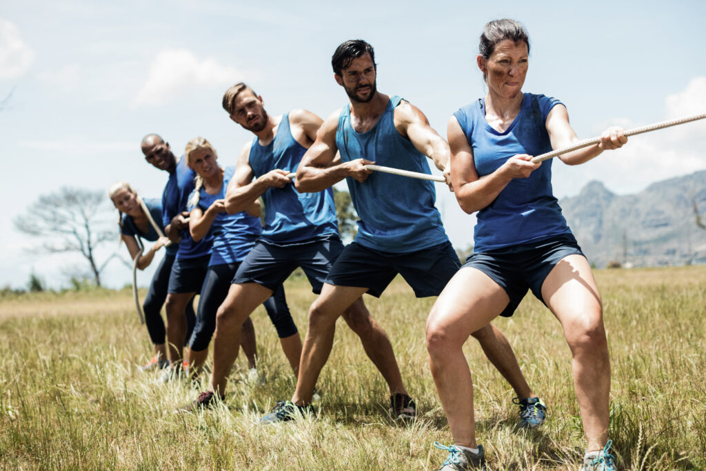 A group of six people in blue athletic gear stand in a line on a grassy field, gripping a thick rope with both hands, leaning back in coordinated effort as they pull together during an outdoor training exercise.