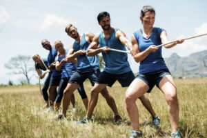 A group of six people in blue athletic gear stand in a line on a grassy field, gripping a thick rope with both hands, leaning back in coordinated effort as they pull together during an outdoor training exercise.