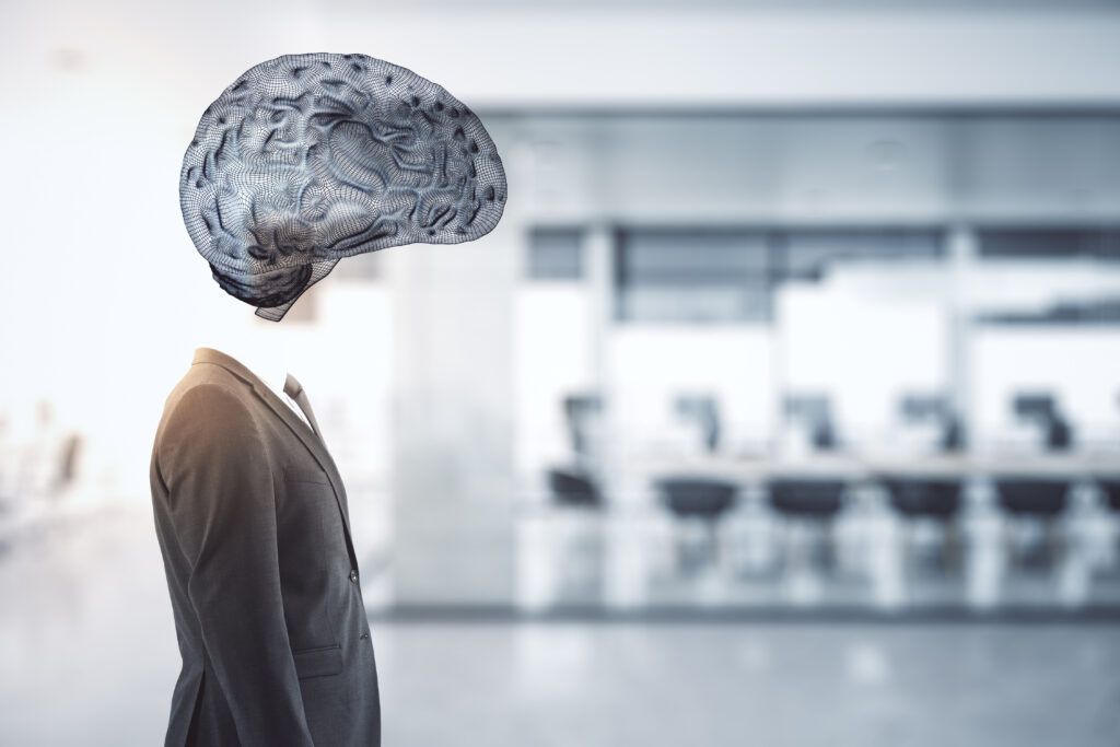 A businessman in a grey suit stands in a blurred office, with a translucent wireframe illustration of a brain in place of his head.