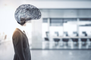 A businessman in a grey suit stands in a blurred office, with a translucent wireframe illustration of a brain in place of his head.