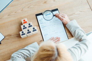 Overhead view of a person in a grey suit examining a printed resume form on a clipboard with a magnifying glass, next to a pyramid of wooden blocks printed with person icons, one of them red.