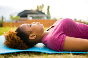 A woman with curly hair lies on her back on a blue yoga mat in the grass outdoors, eyes closed and face relaxed, resting in the late-day light.